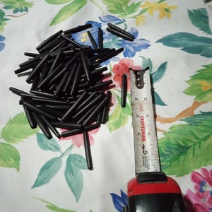 May include: A pile of small, black, tapered plastic tubes. A Craftsman measuring tape shows the tubes are approximately 1 inch long. The background is a floral patterned fabric with blue, yellow, and pink flowers.
