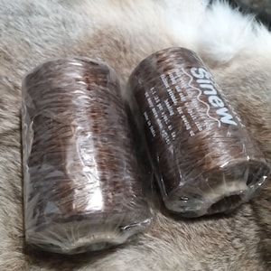 May include: Two spools of brown Sinew thread, wrapped in clear plastic. The text "Sinew" is visible on one spool, along with product details. The spools are resting on a textured, light brown surface.
