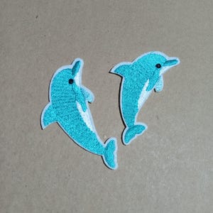 May include: Two embroidered iron-on patches shaped like dolphins. Each dolphin is bright turquoise with a white underbelly and a small black eye. The patches have a white border and are depicted in a leaping pose, showing detailed stitching.