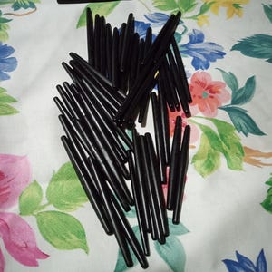 May include: A collection of black stylus pen tips scattered on a floral patterned fabric. The tips are cylindrical with a pointed end, and vary slightly in length. The fabric background features a white base with colorful flowers and green leaves.