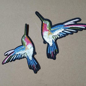 May include: Two embroidered hummingbird patches featuring vibrant thread colors including pink, blue, white, and green. Each bird is depicted in a flying position with wings extended, detailed stitching, and a black border outlining the entire shape.