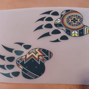 May include: Two black bear paw prints with a colorful geometric design in the center of each print. The design features red, orange, yellow, and black colors.