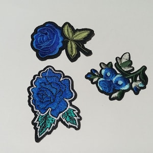 May include: Three embroidered fabric patches featuring floral designs. One patch shows a single blue rose with green leaves, another displays a larger blue rose with teal leaves, and the third depicts a cluster of small blue flowers with green stems.