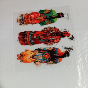 May include: Three colorful Native American Kachina doll stickers. The stickers feature detailed illustrations of the dolls in traditional attire, with vibrant colors including red, green, orange, and black. The stickers are on a clear backing.