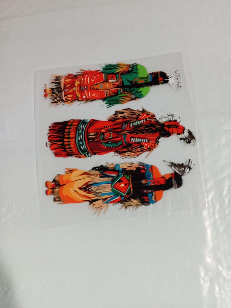 May include: Three colorful Native American Kachina doll stickers. The stickers feature detailed illustrations of the dolls in traditional attire, with vibrant colors including red, green, orange, and black. The stickers are on a clear backing.