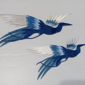 May include: Two embroidered birds in flight, featuring a vibrant blue and white color scheme. The birds have detailed wings and long tail feathers, set against a light background. The design is likely for decorative purposes.