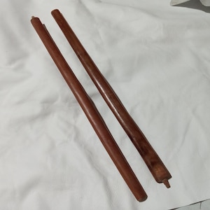 May include: Two wooden furniture legs, each a rich brown color, are positioned diagonally across the frame. The legs are cylindrical, with a slightly tapered design. One leg has a small, square peg at the end. The background is a white, textured surface.