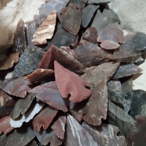 May include: A collection of hand-knapped stone arrowheads in various shades of red, gray, and brown. The arrowheads are of different sizes and shapes, with sharp points and notched bases. The stone appears to be a mix of materials, including flint and chert.