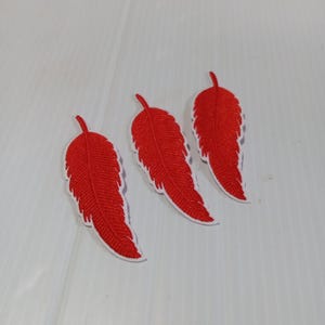 May include: Three red feather patches with white trim are arranged on a white surface. The feathers have a detailed embroidered texture and a small stem at the top. These patches are suitable for clothing or craft projects.