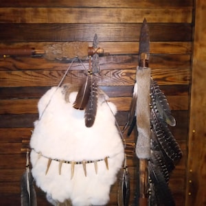 May include: A white rabbit fur hide with a beaded trim and antler accents hangs on a wooden wall with a spear and a feathered staff.