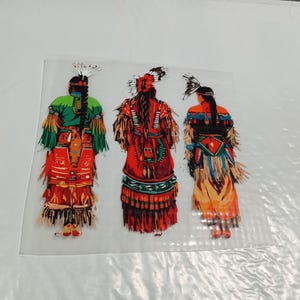 May include: Three figures in traditional Native American attire are depicted from the back. The figures are adorned in vibrant colors, including red, green, and orange, with intricate details in their clothing and headdresses. The image is set against a white background.