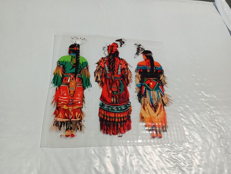 May include: Three figures in traditional Native American attire are depicted from the back. The figures are adorned in vibrant colors, including red, green, and orange, with intricate details in their clothing and headdresses. The image is set against a white background.