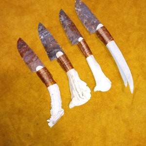 May include: Four handmade knives with brown leather handles and white bone handles. The blades are made of a dark grey stone.
