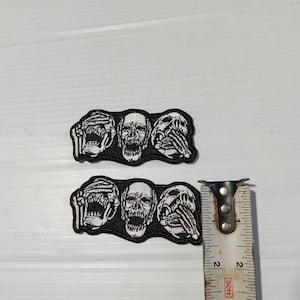 May include: Two black and white embroidered patches. Each patch features three skulls, one covering its eyes, one with an open mouth, and one covering its mouth. A measuring tape is next to the patches.
