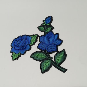 May include: Two embroidered patches featuring vibrant blue roses with bright green leaves. One patch shows a single rose bloom with foliage, while the other depicts a rose stem with a bud, a full bloom, and several leaves, all outlined in black.