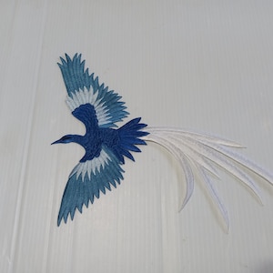 May include: Embroidered patch featuring a stylized bird in flight. The bird has a dark blue body, wings with shades of blue and white, and long white tail feathers. The patch is likely for decorative purposes.