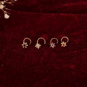 Peut inclure: Quatre piercings de nez dorés avec des motifs floraux. Chaque bijou présente une petite fleur ornée de pierres précieuses de différentes couleurs : vert, transparent, vert et rose. Les anneaux sont présentés sur un fond de velours rouge foncé.