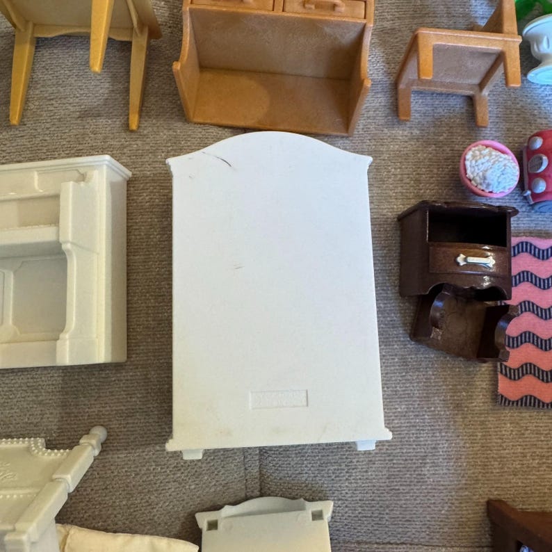 May include: A collection of miniature dollhouse furniture pieces including a white wardrobe, a brown nightstand with a drawer, a tan bench, a white shelf unit, a small pink bowl with white contents, and a pink and black patterned rug piece.