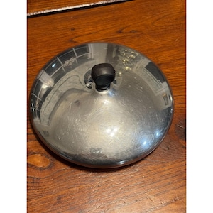 May include: A shiny, stainless steel lid with a black knob. The lid is round and reflects the surrounding environment. The knob is small and cylindrical. The lid is sitting on a wooden surface.
