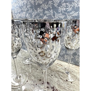 May include: Set of clear wine glasses with silver rims and a textured, leopard-print-like pattern. The glasses have long stems and wide bowls, reflecting light. The background features a floral pattern.