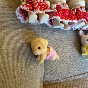May include: A collection of small flocked animal figurines including a crawling puppy wearing a pink diaper and several standing figures dressed in red dresses with white polka dots and white lace trim, alongside a figure in a yellow dress.