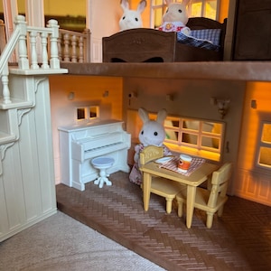 May include: A multi-level dollhouse interior featuring a white upright piano with a matching stool, a tan dining table with two chairs, and three white rabbit figurines. The scene includes a brown bed frame and a staircase with white railings.
