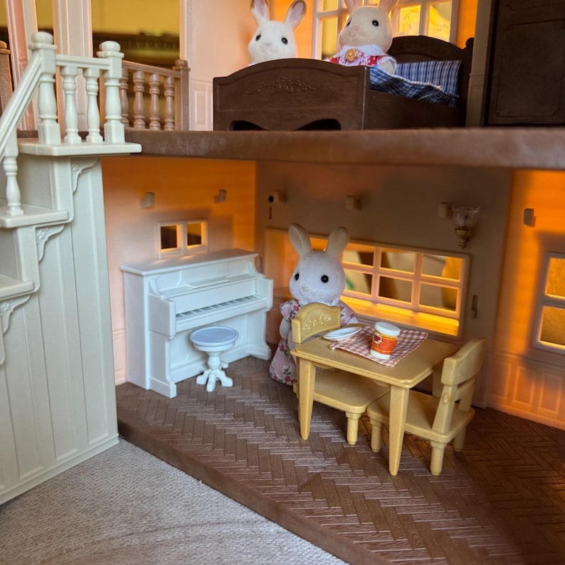 May include: A multi-level dollhouse interior featuring a white upright piano with a matching stool, a tan dining table with two chairs, and three white rabbit figurines. The scene includes a brown bed frame and a staircase with white railings.
