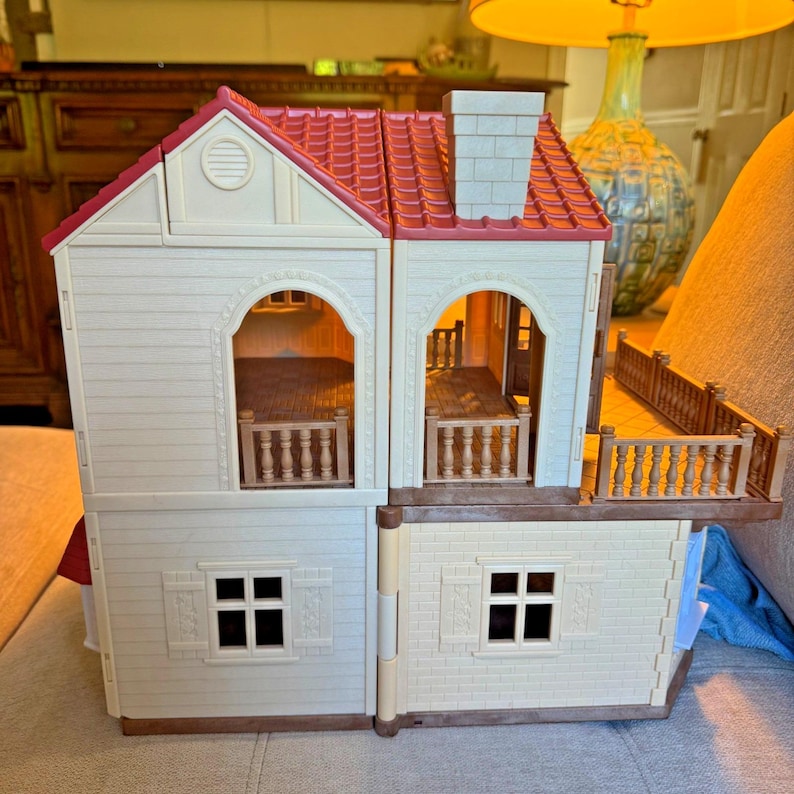 May include: A two-story toy house with cream-colored walls, a red tiled roof, and a chimney. The structure features arched doorways, brown railings on the balconies, and windows with shutters on the lower level. The house is open-faced.