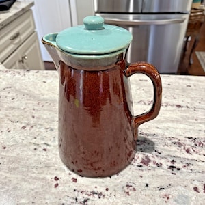 May include: A brown ceramic pitcher with a teal lid and handle. The pitcher has a glossy finish and a classic design, suitable for serving beverages. The handle is a darker shade of brown, complementing the overall color scheme.