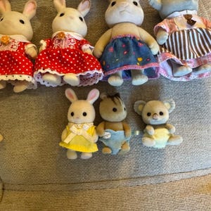May include: A collection of seven miniature flocked animal figurines. The group includes four rabbits in dresses, one bear in a blue diaper, one koala in a yellow diaper, and one mouse wearing a pink and brown striped dress with a white collar.