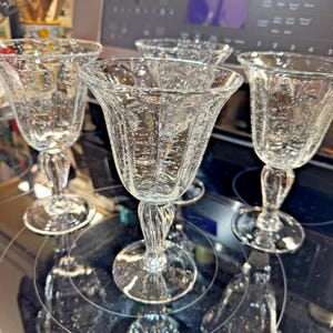 May include: Set of four clear, stemmed wine glasses. Each glass features a textured, bubble-like design throughout the bowl and a decorative stem. The glasses are arranged on a reflective black surface.