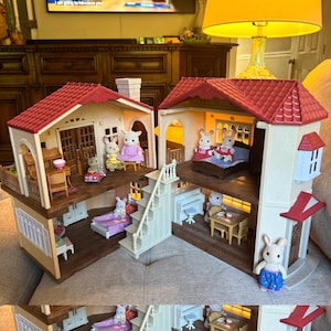May include: A two-story dollhouse with a red shingled roof, cream-colored walls, and brown floors. The interior features multiple rooms furnished with miniature tables, chairs, beds, and a piano, populated by several small flocked rabbit figurines.