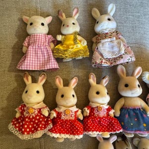 May include: A collection of seven flocked animal figurines, primarily rabbits, wearing various outfits including a pink gingham dress, a yellow dress, a floral dress with a white apron, and several red dresses with white polka dots and lace trim.