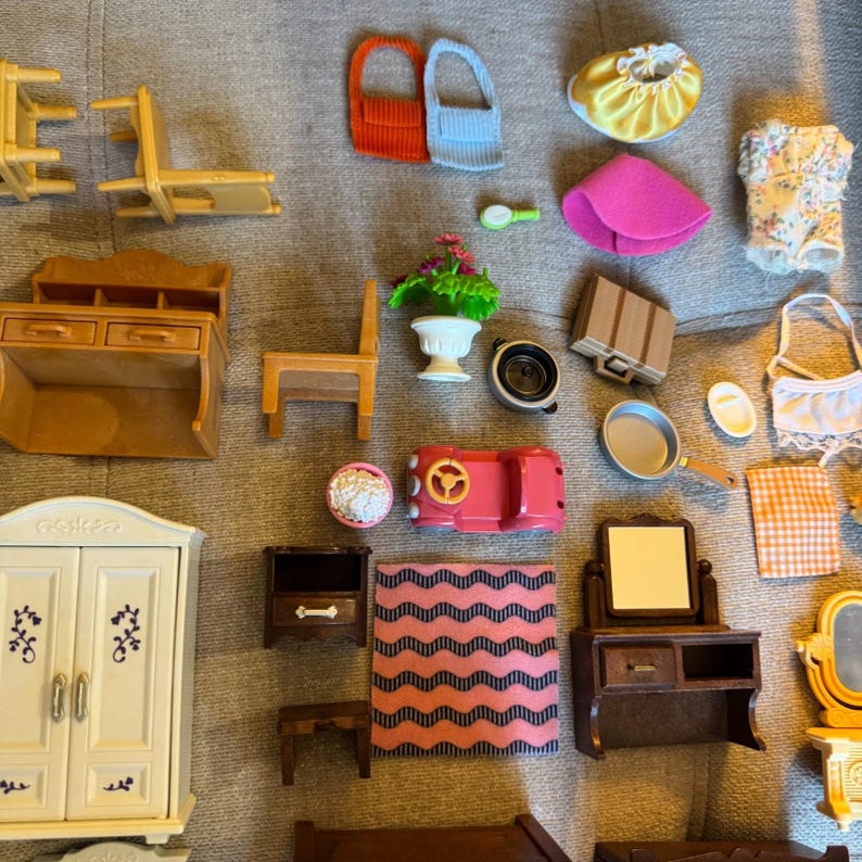 May include: A collection of miniature dollhouse furniture and accessories including a white wardrobe, brown vanity, wooden chairs, a pink toy car, a frying pan, a potted plant, various fabric clothing items, a small rug, and assorted household trinkets.