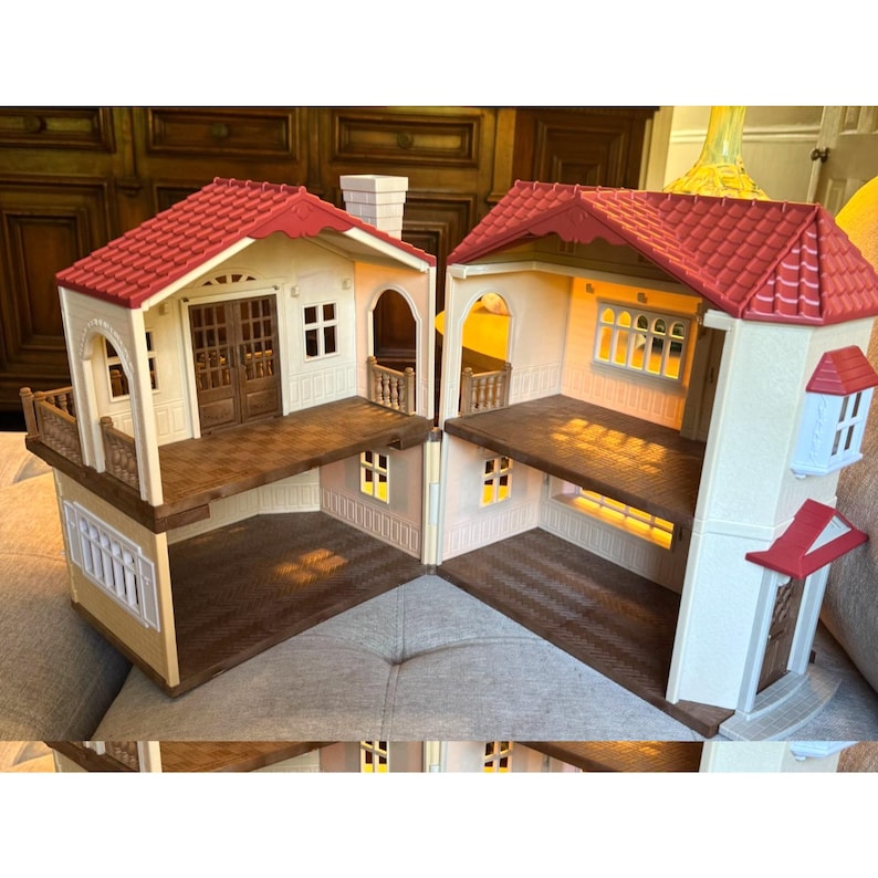 May include: A two-story dollhouse with cream-colored walls and a red tiled roof. The structure features brown floors, a balcony with railings, multiple windows with white frames, and a set of brown double doors. The interior is open-plan.