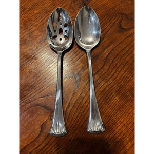 May include: Two silver serving spoons on a wooden surface. One spoon has a slotted bowl, and the other has a solid bowl. Both spoons have long, ribbed handles and a decorative base. The spoons are likely for serving food.