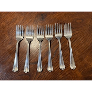May include: Seven silver forks with tines and long handles are arranged on a dark wooden surface. The forks are made of a shiny metal and have a classic design, suitable for formal dining.