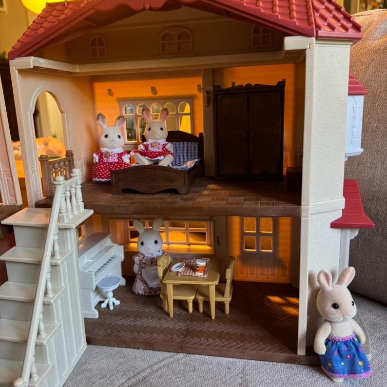 May include: A two-story dollhouse with a red roof, featuring a bedroom on the top floor with a bed and wardrobe, and a living area on the bottom floor with a piano, table, and chairs. Five rabbit figurines in various outfits are positioned throughout.