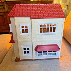 May include: A two-story dollhouse with cream-colored walls and a textured red roof. The structure features multiple white-framed windows, including a large bay window on the ground floor with a red awning and a wide window on the second floor.