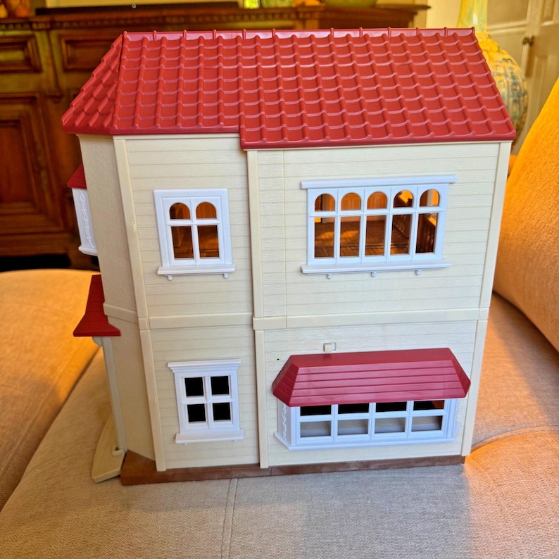 May include: A two-story dollhouse with cream-colored walls and a textured red roof. The structure features multiple white-framed windows, including a large bay window on the ground floor with a red awning and a wide window on the second floor.