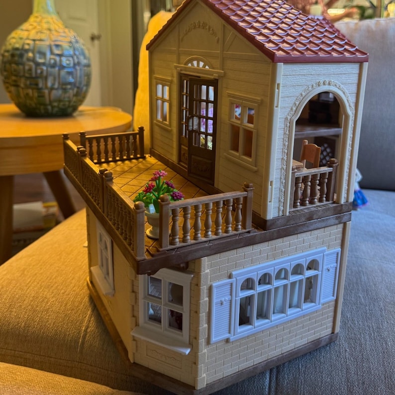 May include: A two-story toy house with a red shingled roof, cream-colored walls, and a brown balcony featuring a small potted plant with pink flowers. The structure includes multiple windows with white frames, a brown front door, and a side archway.