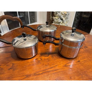 May include: Three stainless steel pots with lids and black handles are arranged on a wooden table. The pots vary in size, with a double-tiered pot on the right. The lids have black knobs.