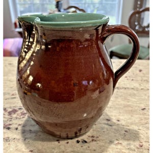May include: A handmade ceramic pitcher with a dark brown exterior and a teal green interior rim. The pitcher has a curved handle and a spout for pouring. The glaze gives the pitcher a glossy finish. The pitcher is likely used for serving beverages.