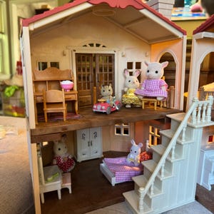 May include: A two-story dollhouse with a red roof, featuring a desk, chair, and pink bowl on the top floor alongside three animal figures, one in a pink car. The bottom floor contains a bed with a purple checkered blanket, a white cabinet, and two figures.