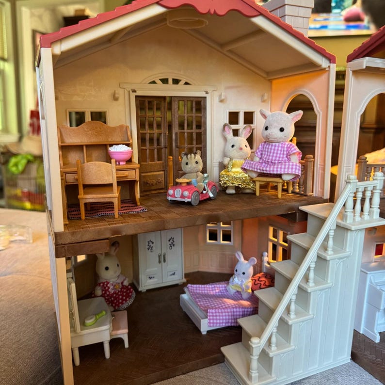 May include: A two-story dollhouse with a red roof, featuring a desk, chair, and pink bowl on the top floor alongside three animal figures, one in a pink car. The bottom floor contains a bed with a purple checkered blanket, a white cabinet, and two figures.