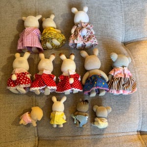 May include: A collection of thirteen miniature flocked animal figurines, including rabbits, koalas, and other creatures. The figures are shown from the back, wearing various colorful fabric outfits such as dresses with floral, polka dot, and plaid patterns.