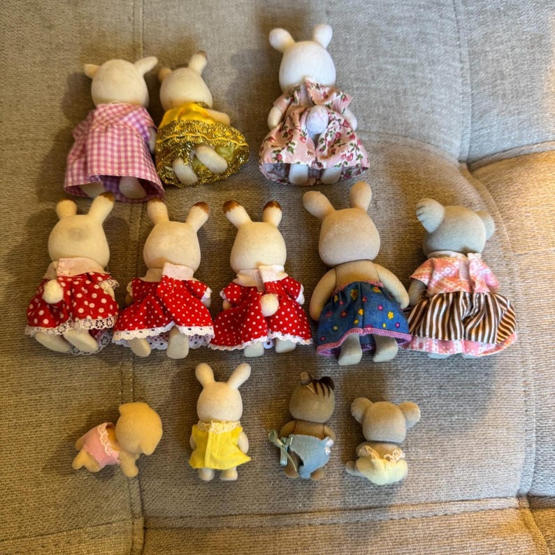 May include: A collection of thirteen miniature flocked animal figurines, including rabbits, koalas, and other creatures. The figures are shown from the back, wearing various colorful fabric outfits such as dresses with floral, polka dot, and plaid patterns.