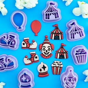 Puede incluir: Una colección de cortadores de galletas de plástico morado y adornos de fondant a juego con temas de circo, incluyendo una cara de payaso, carpas a rayas, cubos de palomitas, globos y zapatos grandes sobre un fondo azul.