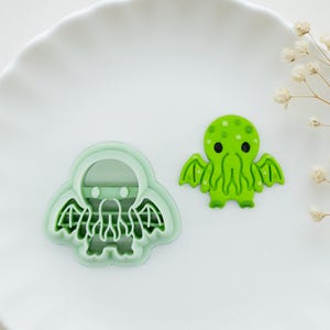 May include: A green Cthulhu-shaped cookie cutter and a green Cthulhu-shaped decoration. The cookie cutter is a light green color and has a detailed design. The decoration is a brighter green with black eyes.