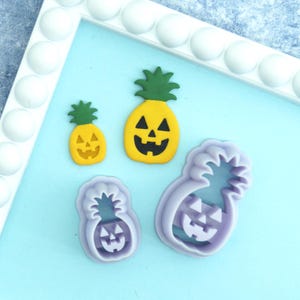 May include: Two yellow pineapple-shaped cutouts with green tops and black jack-o'-lantern facial features, paired with two matching light purple plastic cookie cutters in different sizes, arranged on a light blue surface.